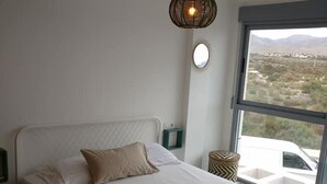 Ett modernt sovrum med en stor säng, en hängande lampa, utsikt över öknen genom ett stort fönster och ett litet sidobord.