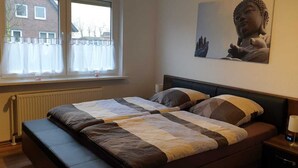Une chambre à coucher avec un lit, une table de chevet, une lampe, une fenêtre avec des rideaux et une statue de Bouddha accrochée au mur.
