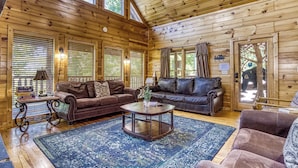 L’intérieur chaleureux d’un chalet en bois comprend deux canapés en cuir, une table basse ronde et une grande fenêtre équipée de stores.