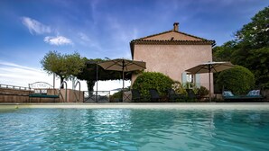 A swimming pool with lounge chairs and umbrellas, a brick building, and greenery in the background. - Restored farmhouse with heated pool and landscaped gardens. (Apt)