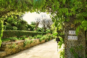 Um caminho de pedra serpenteia por um jardim exuberante, com sebes bem aparadas e uma placa com a inscrição 'CHEZ MANON'.