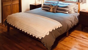 Ein Schlafzimmer mit Holzbalkendecke, einem Bett mit weißer und grauer Bettdecke, zwei Nachttischen mit Lampen und einem Fenster mit Vorhängen.
