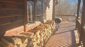 Eine Holzterrasse mit gestapeltem Brennholz, einem Grill und Blick auf Bäume und Berge.