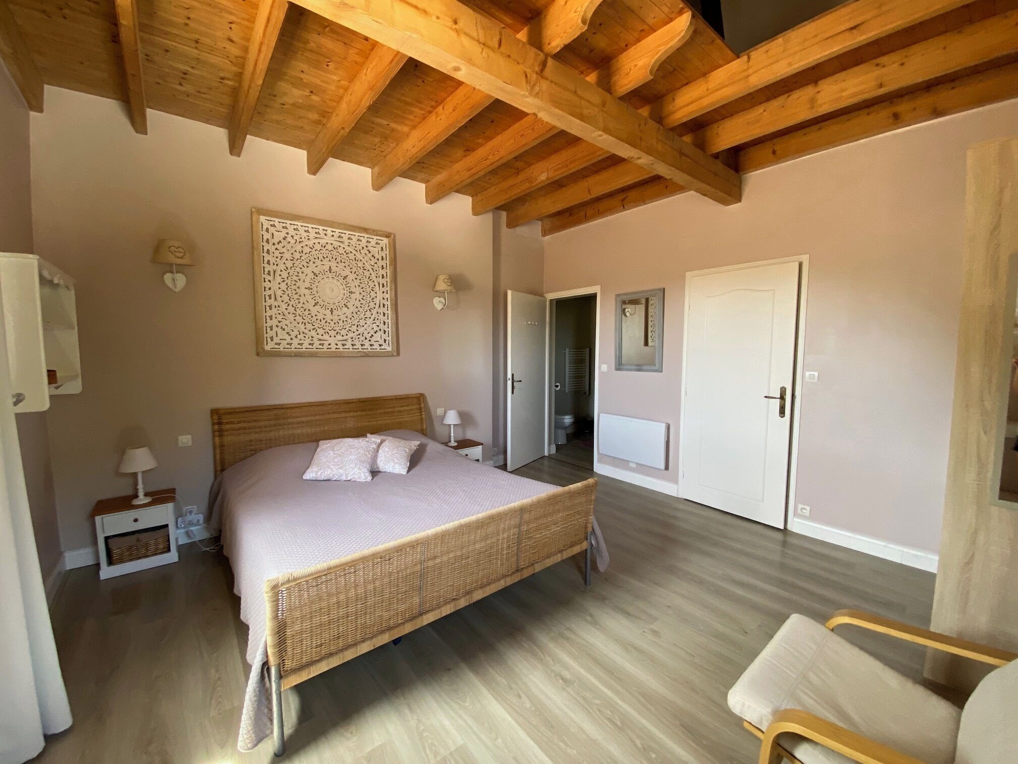 Quarto com cama grande, vigas de madeira no teto, um objeto decorativo na parede, uma porta e uma cadeira.