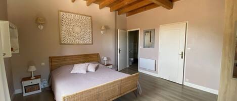 Quarto com cama grande, vigas de madeira no teto, um objeto decorativo na parede, uma porta e uma cadeira.