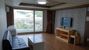 A modern living room with a sofa, a coffee table, a wooden TV stand, and a large sliding glass door leading to a balcony. - Hawaii Hotel (Tongyeong)