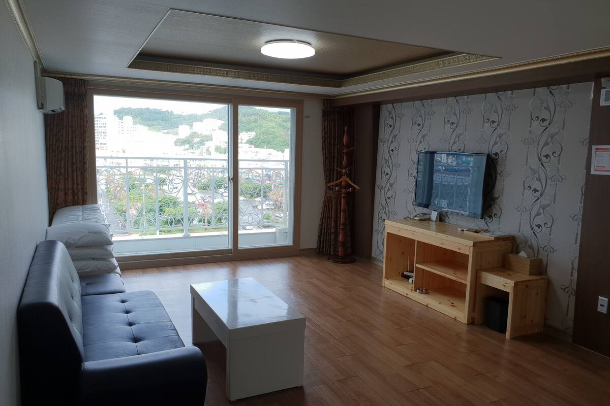 A modern living room with a sofa, a coffee table, a wooden TV stand, and a large sliding glass door leading to a balcony.