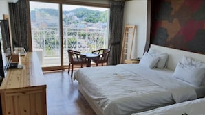 A hotel room with a large bed, a desk, a chair, and a balcony with a view of the city. - Hawaii Hotel (Tongyeong)