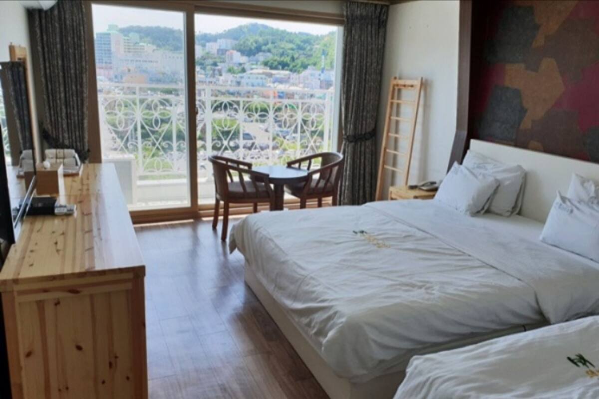 A hotel room with a large bed, a desk, a chair, and a balcony with a view of the city.