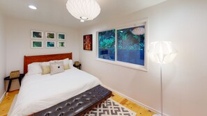 Une chambre avec un grand lit, une tête de lit en bois, un tapis à motifs, une fenêtre donnant sur un paysage verdoyant et un luminaire fixé au mur.