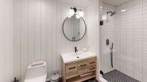 Une salle de bain moderne avec un meuble-lavabo blanc, un grand miroir rond doté de lumières, des toilettes et un espace douche avec une porte en verre.
