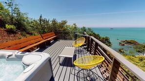 Une terrasse en bois avec un jacuzzi, deux chaises jaunes et un banc en bois, le tout donnant sur une mer d’un bleu limpide.