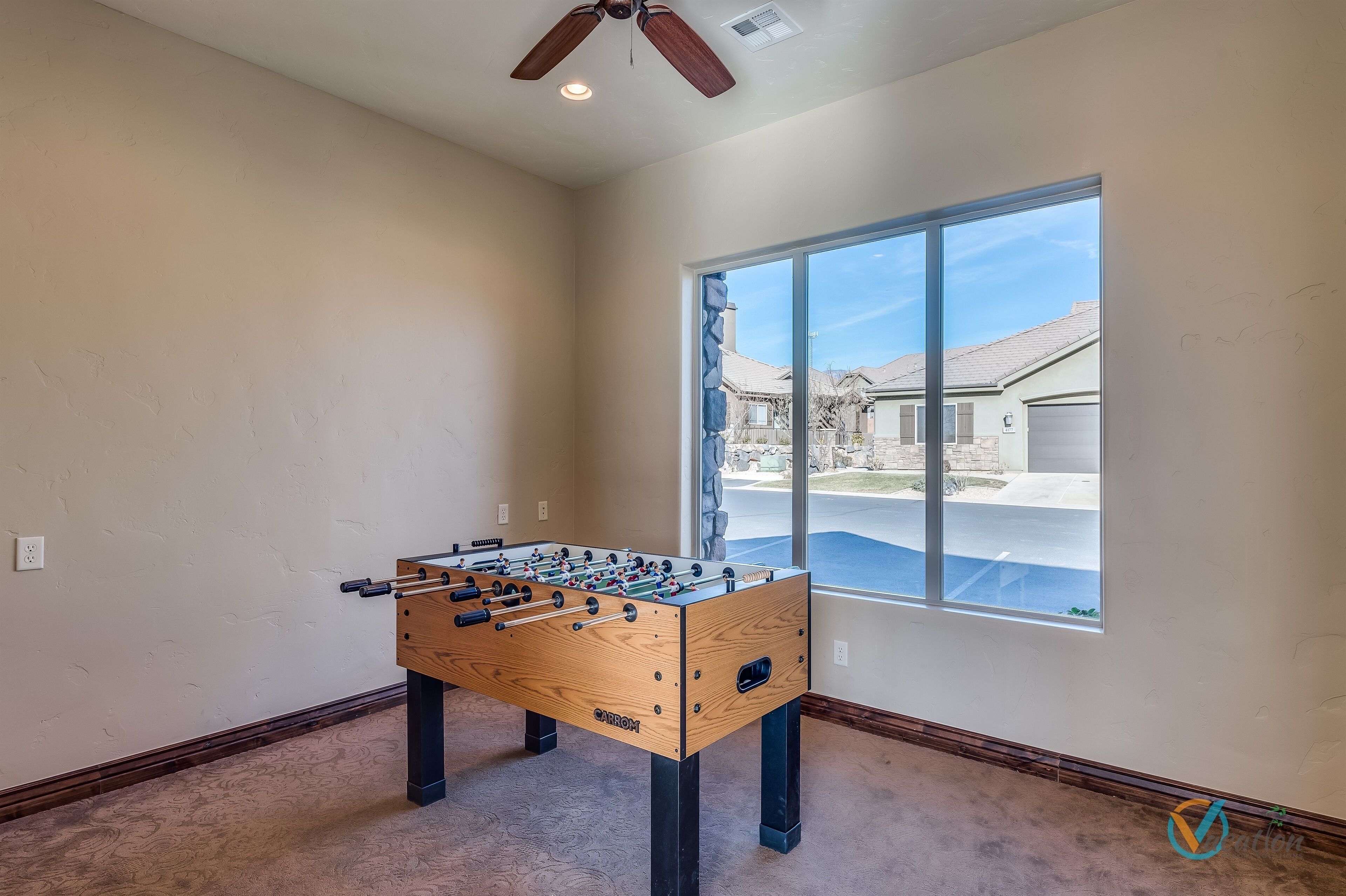 A room with a foosball table, a ceiling fan, and a large window offering a view of a suburban neighborhood.