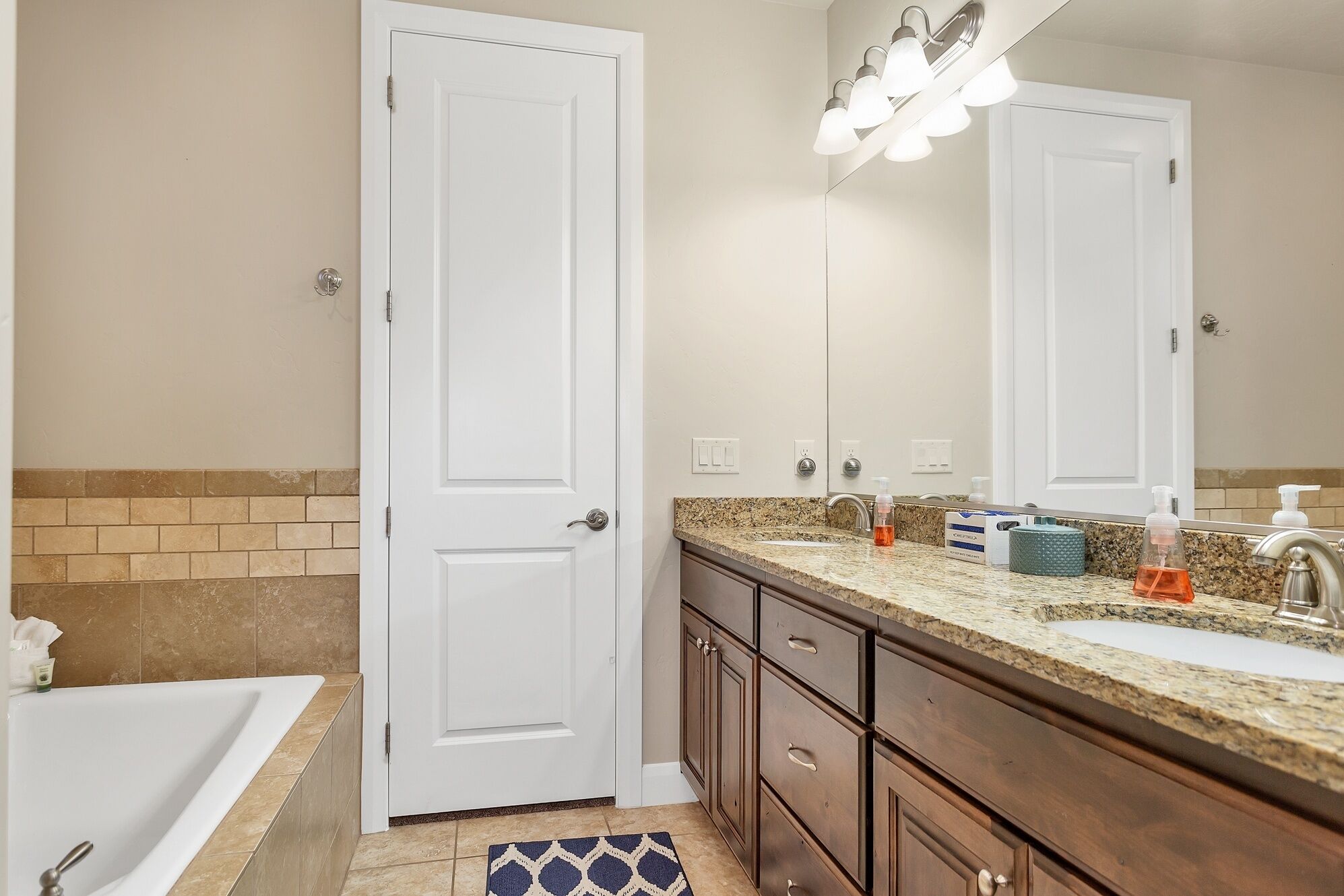 A bathroom with a white bathtub, a sink with a granite countertop, and a mirror above the sink. There are toiletries on the countertop and a towel rack on the wall.