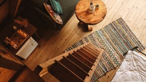 Una habitación con piso de madera, un sofá verde, una mesa de centro redonda de madera, un tapete estampado y una cama con sábanas blancas.