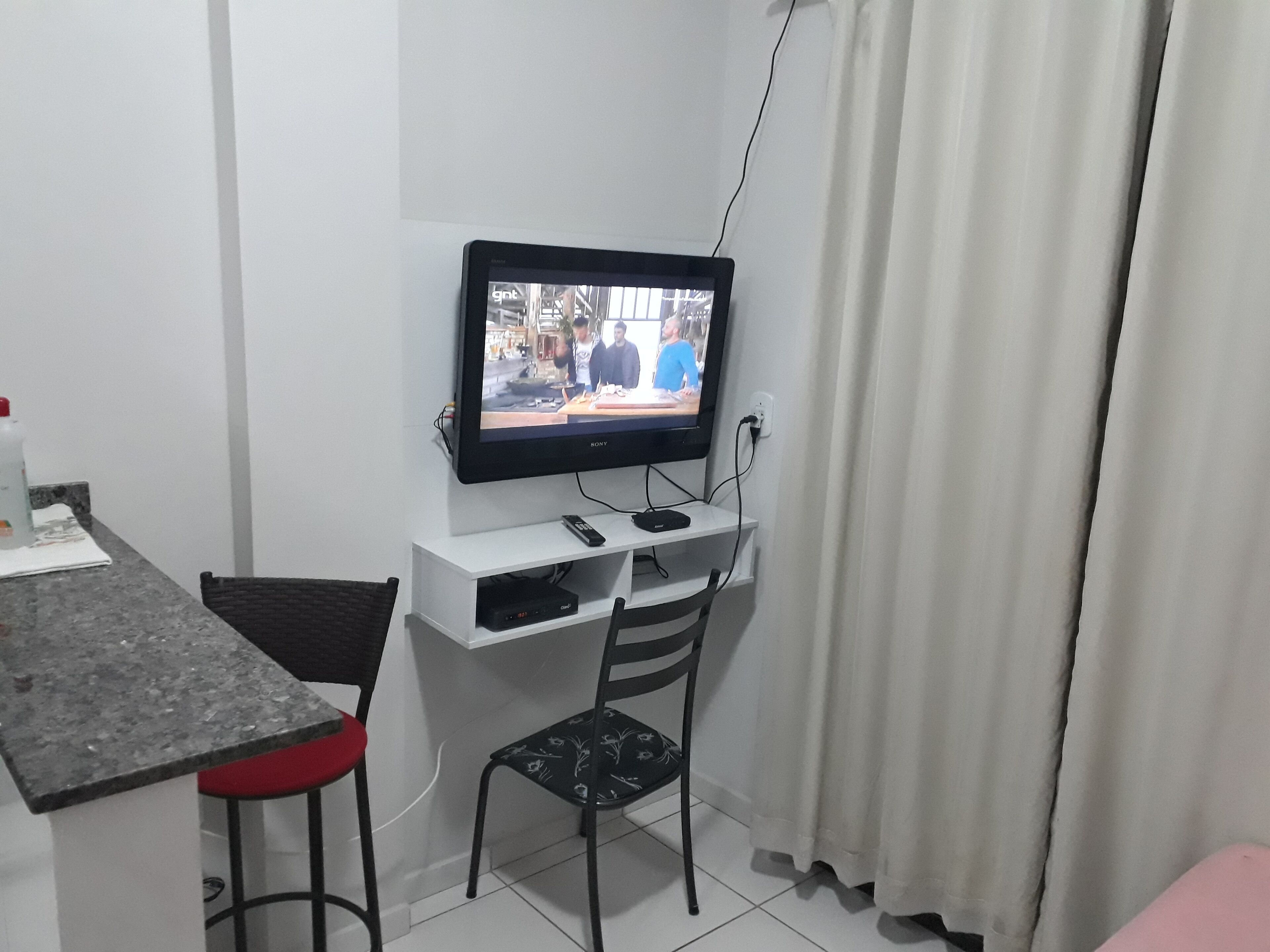 Ett litet vardagsrum med en tv monterad på en vit vägg, ett litet bord med en röd stol och en svart stol med ett blommönster.