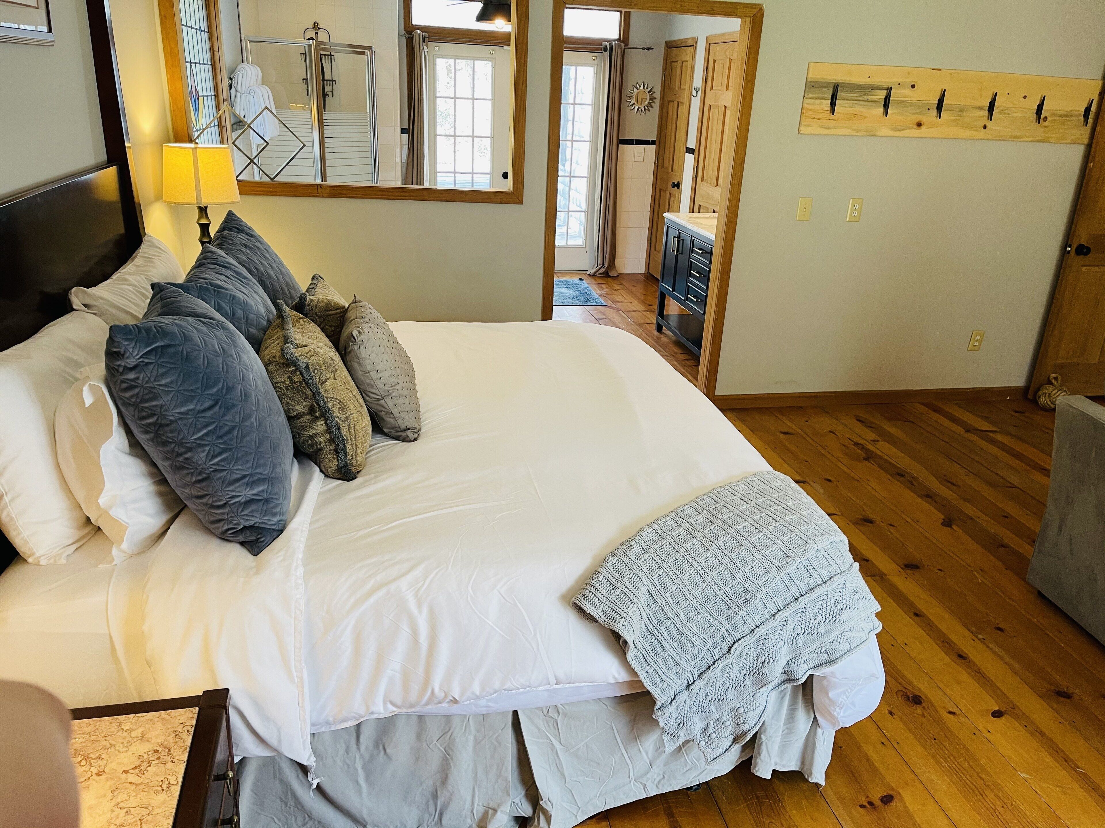 A bedroom with a large bed, multiple pillows, a bedside lamp, a wooden floor, a mirror, and a door leading to another room.