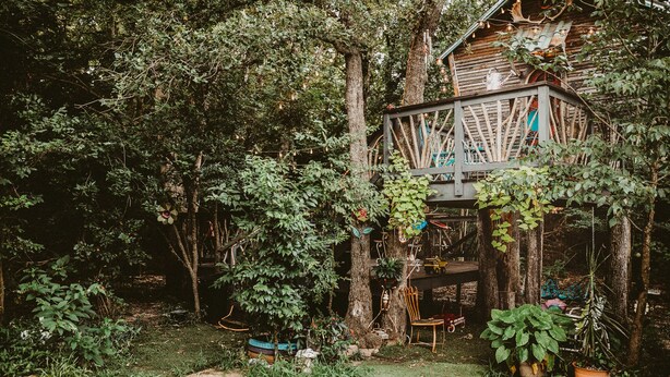 A rustic wooden treehouse with a wooden deck, surrounded by lush greenery and potted plants. - The Treehouse, with a deck for grilling and relaxation. (Terrell)