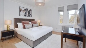 A modern bedroom with a large bed, bedside tables, a desk, a TV, and artwork on the wall. - 3 bedroom apartment in the heart of Kensington (London)
