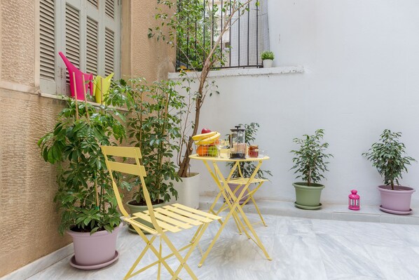 A small outdoor seating area with a table and chairs, surrounded by potted plants and a window with shutters.