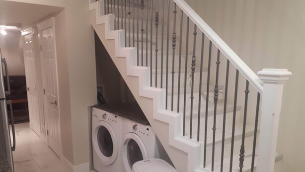 A laundry area with two washing machines and a trash bin, a staircase with white railings, and a door leading to another room.
