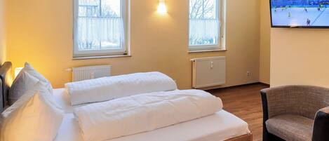 Habitación de hotel con cama, dos ventanas, un radiador, un televisor y una silla.