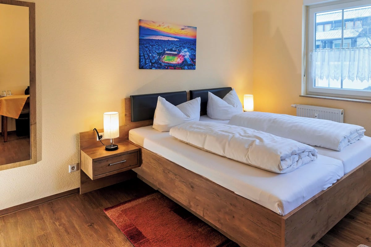 A hotel room with a wooden bed, white bedding, a nightstand with a lamp, a red rug, a painting of a stadium, and a window with a view of buildings.