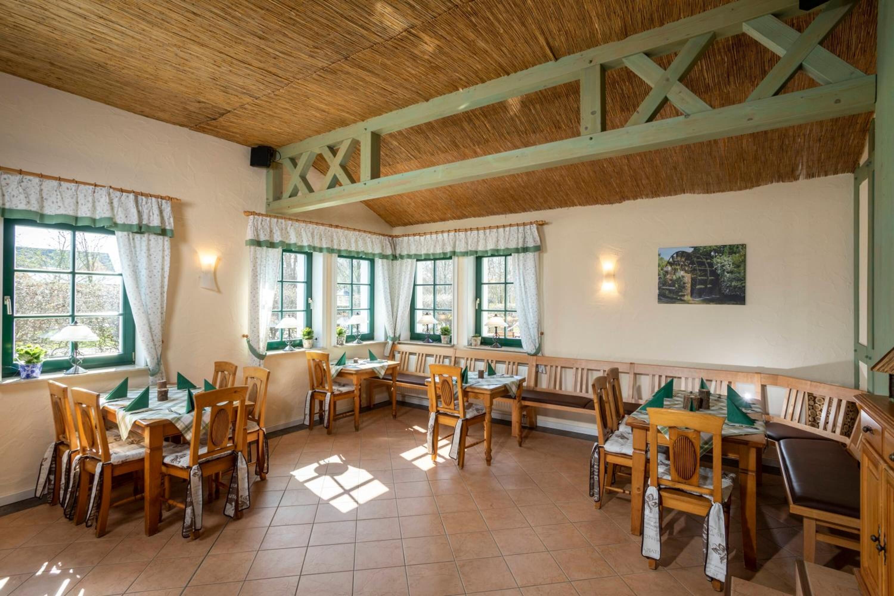 A dining area with wooden tables and chairs, large windows with green curtains, and a thatched roof ceiling.