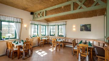Uma sala de jantar com mesas e cadeiras de madeira, grandes janelas com cortinas verdes e teto de colmo.