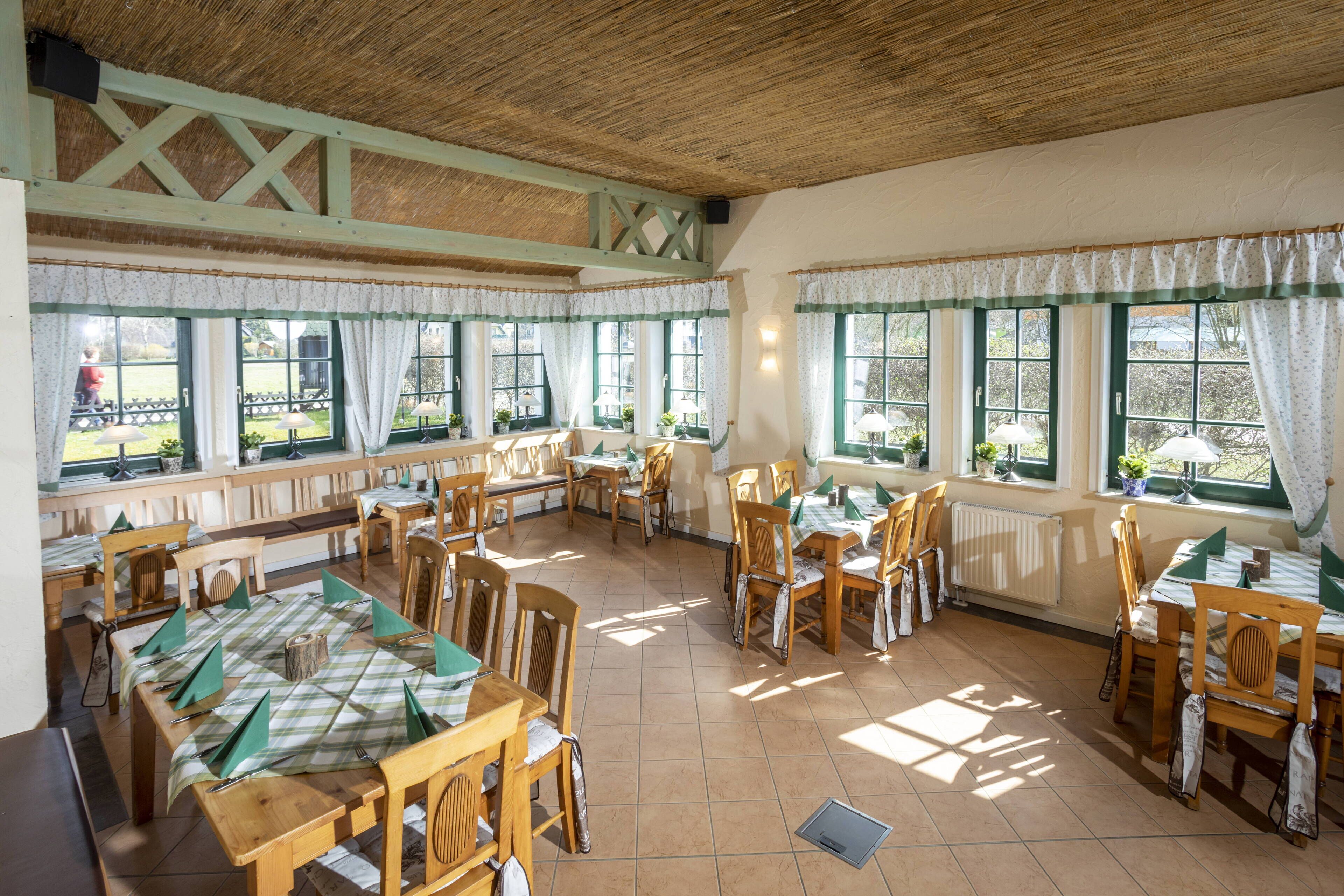 A dining area with wooden tables and chairs, large windows with curtains, and a rustic ceiling.