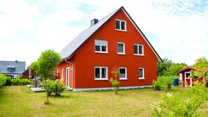 Une maison rouge à deux étages, avec un toit en pente, entourée d’une pelouse bien entretenue et de petits arbres.