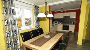 Un coin repas avec une table et des chaises en bois, une fenêtre avec des rideaux et, au fond, une cuisine.