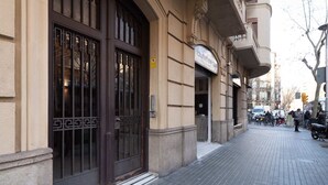A building with a large dark wooden door and a sign above it. There are bicycles parked outside and a few people walking. - Sweett | Sepulveda Principal A (Barcelona)
