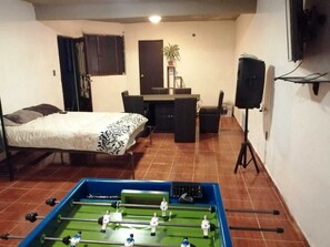 A room with a bed, a desk, a chair, a foosball table, and a TV.