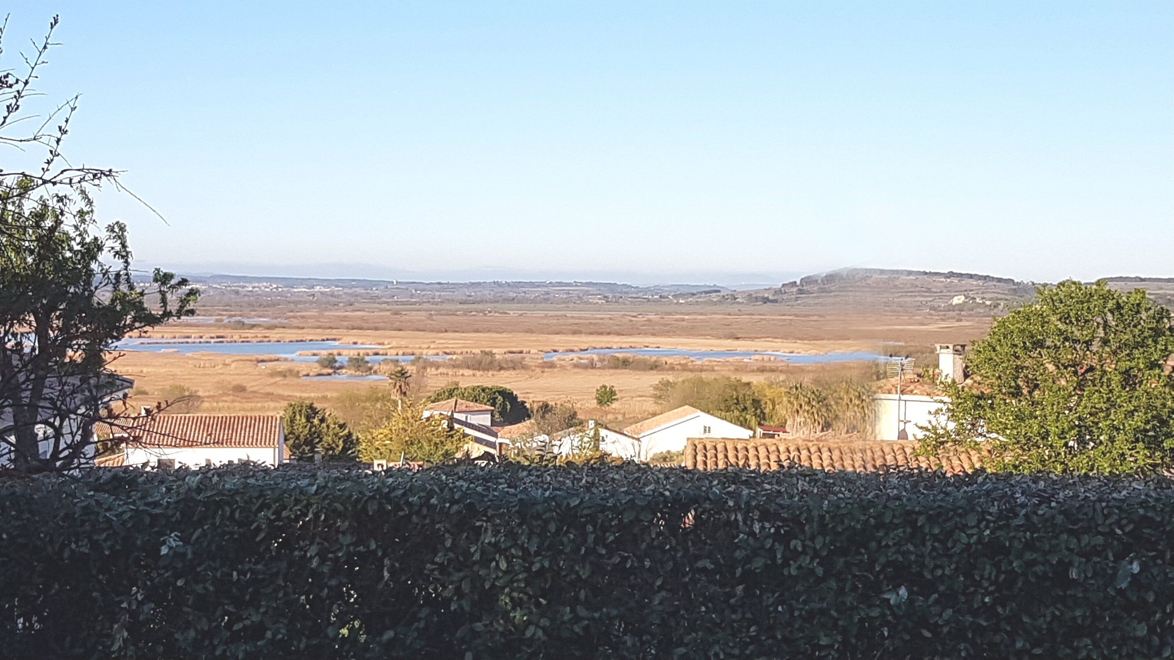 Paisagem rural com casas, um rio e colinas ao longe.