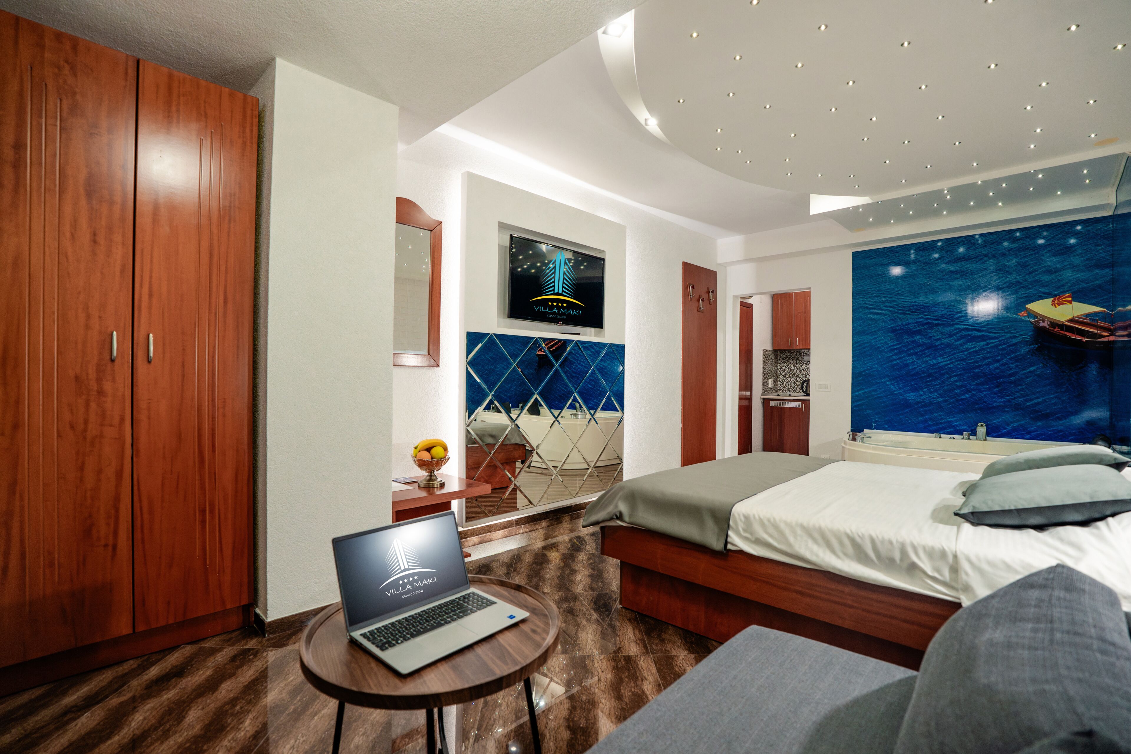 A hotel room with a bed, a laptop on a small table, a TV mounted on the wall, and a large window with a view of the sea.
