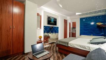A hotel room with a bed, a laptop on a small table, a TV mounted on the wall, and a large window with a view of the sea.