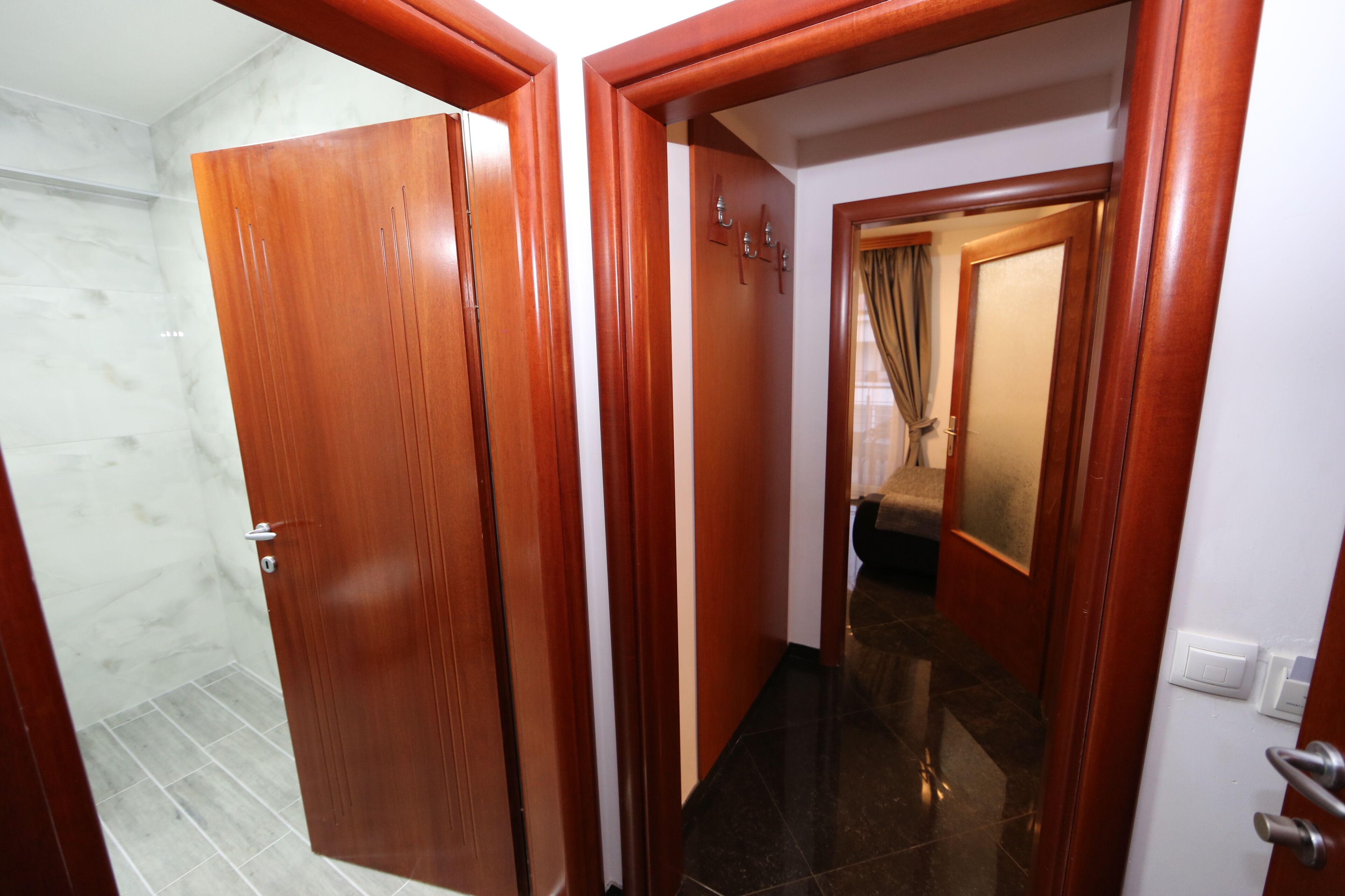 Un couloir avec des portes en bois, un sol carrelé et un miroir qui reflète une pièce avec un lit et des rideaux.