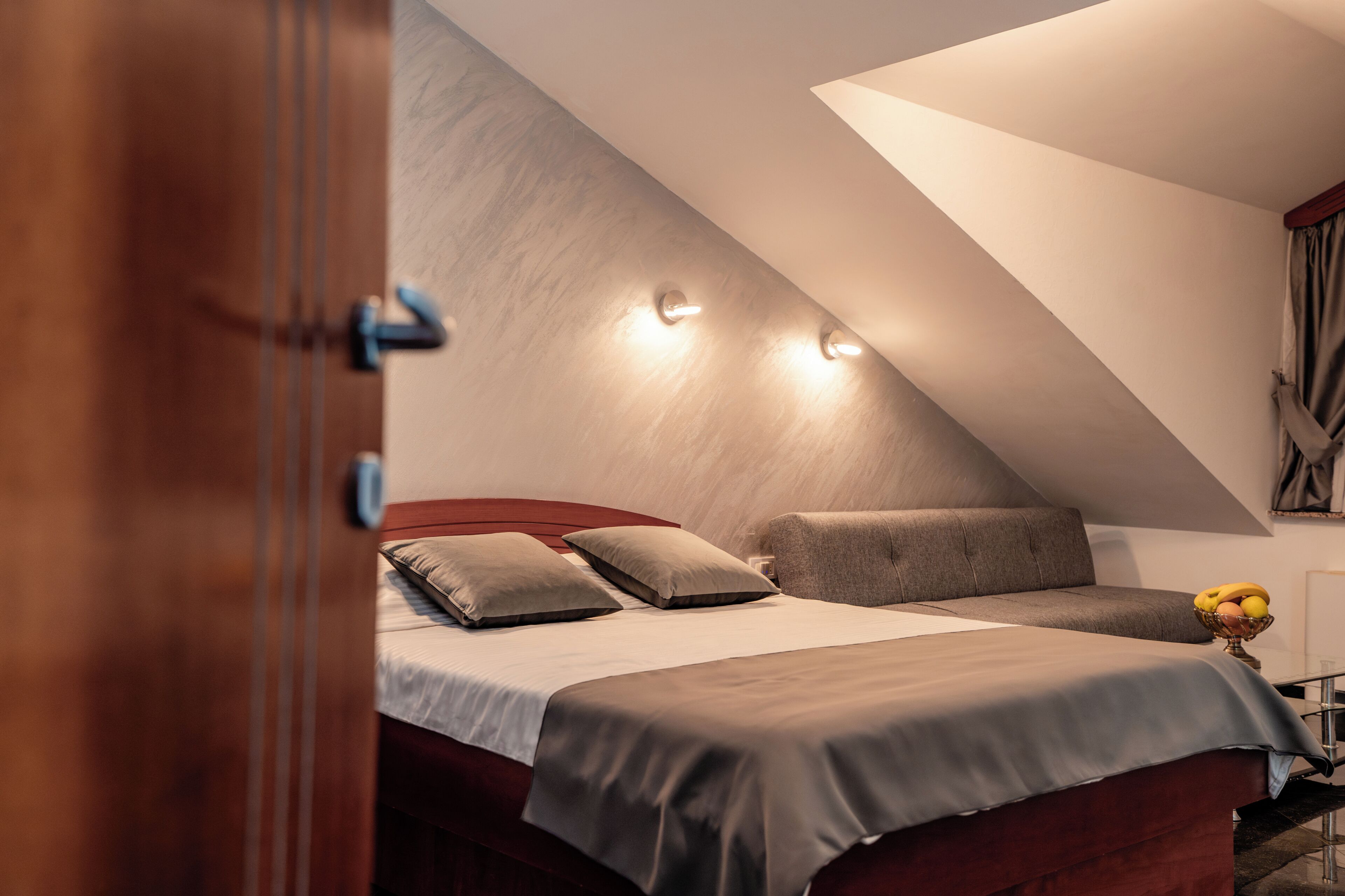 A bedroom with a sloped ceiling, a bed with two pillows, a bedside table with a fruit bowl, and a wall-mounted light fixture.