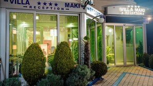 Recepção de hotel com uma placa indicando 'VILLA MAKI RECEPTION', porta de vidro de entrada e um pequeno jardim com arbustos variados.