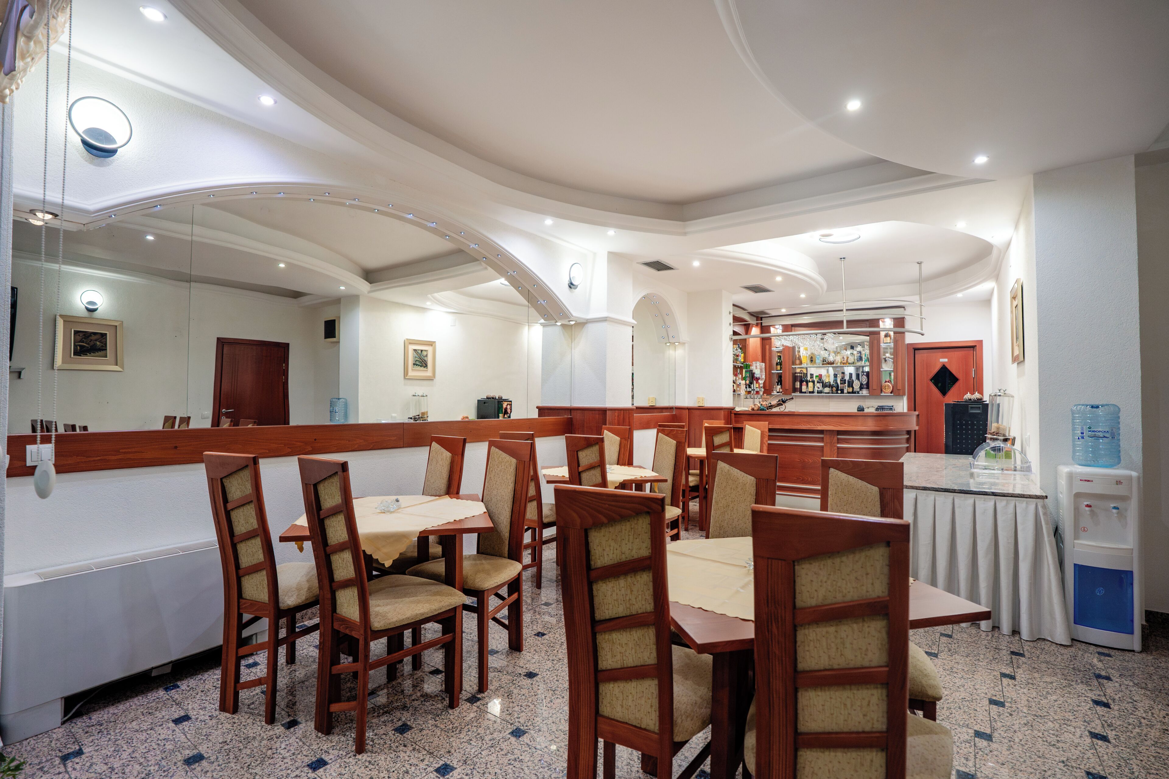A restaurant with wooden chairs and tables, a counter with a display, and a water dispenser.