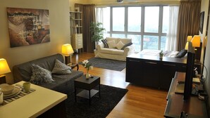 A modern living room with a sofa, coffee table, and a desk with a computer. There is a large window with curtains, a ceiling fan, and a painting on the wall. - Regalia Suites Infinity Pool (Kuala Lumpur)