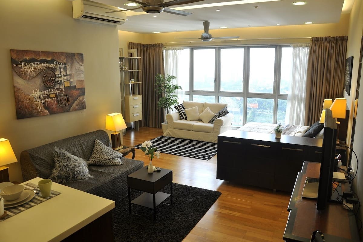 A modern living room with a sofa, coffee table, and a desk with a computer. There is a large window with curtains, a ceiling fan, and a painting on the wall.