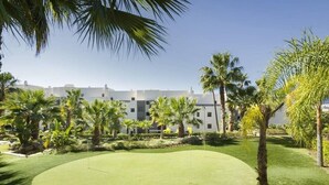 A golf course with a well-maintained green, surrounded by palm trees and a building in the background. - Hoyo19 11 | Premier Luxury Holiday Resort (Málaga)