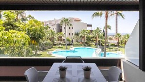 A balcony with a table and chairs, overlooking a swimming pool and a building with palm trees. - Hoyo19 11 | Premier Luxury Holiday Resort (Málaga)