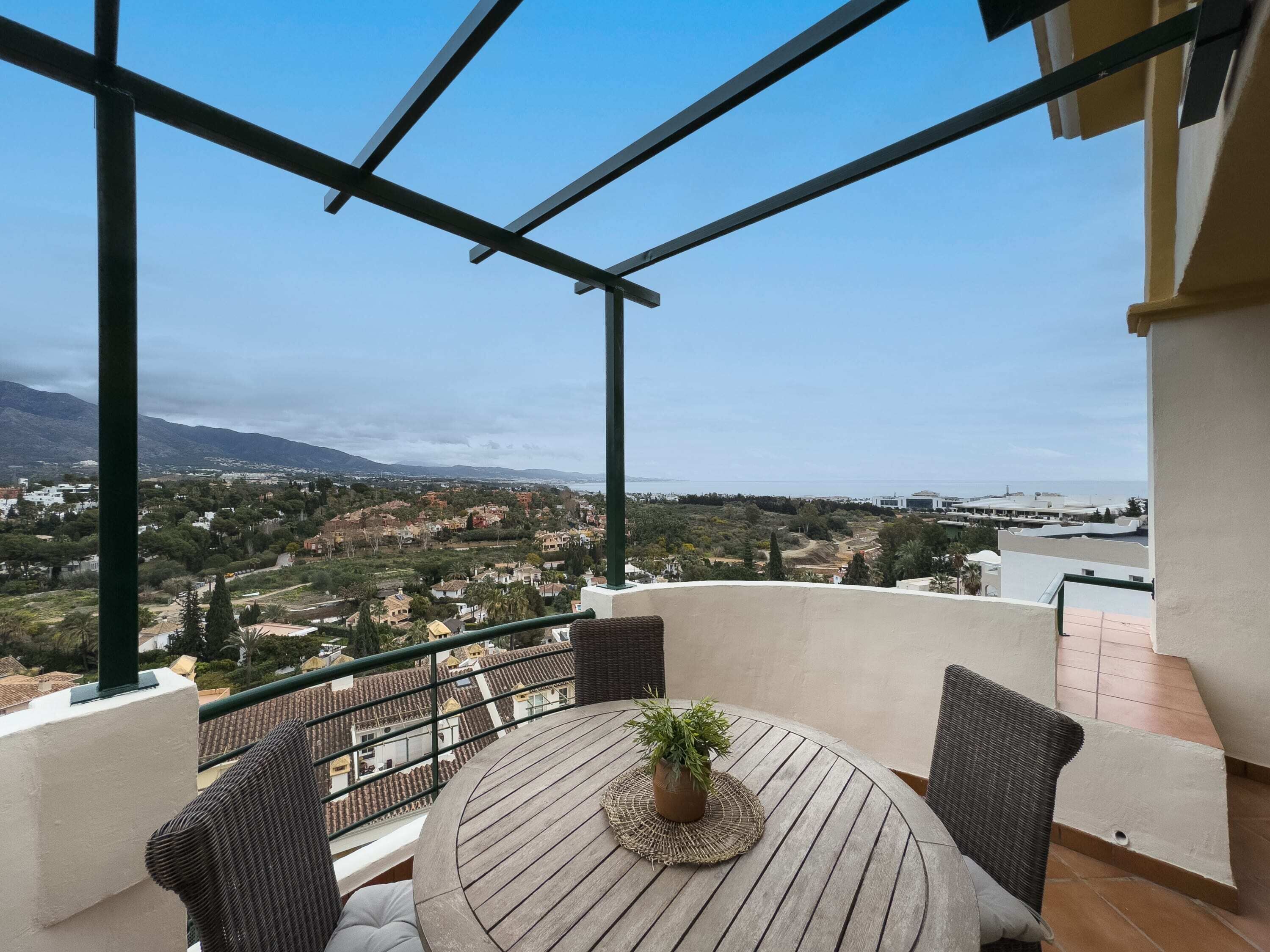 Een balkon met een ronde houten tafel en twee rieten stoelen, met uitzicht op een kustplaatsje met gebouwen en bergen in de verte.