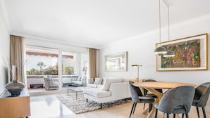 Een moderne woonkamer met een witte bank, een eettafel en stoelen, een groot raam met gordijnen en een schilderij aan de muur.