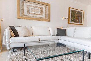 Ein modernes Wohnzimmer mit einem weißen Ecksofa, einem Glastisch, einem flauschigen Teppich und zwei gerahmten Kunstwerken an der Wand.