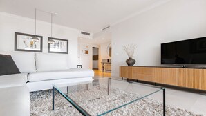 Een moderne woonkamer met een glazen salontafel, een witte bank en een televisie op een houten dressoir.