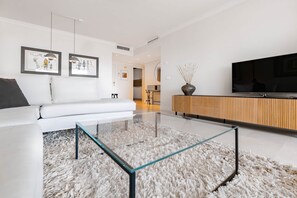Un salon moderne avec une table basse en verre, un canapé blanc et un téléviseur posé sur un meuble en bois.